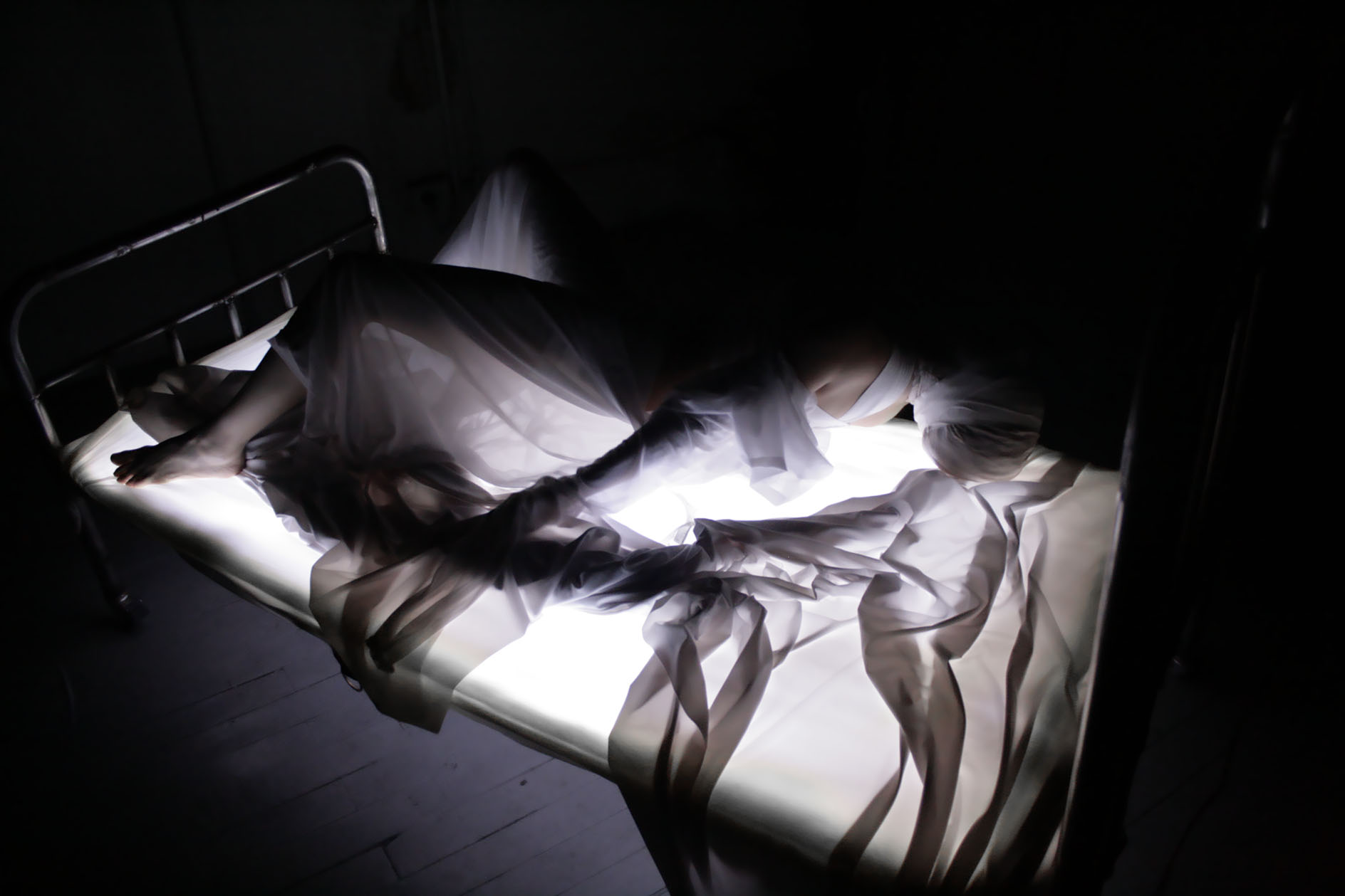 Una bailarina tendida sobre una cama de hierro, cubierta de tul iluminado.